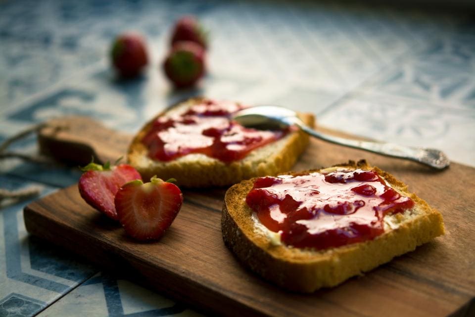 Duas fatias de pão com geleia de morango numa tábua, com morangos ao lado