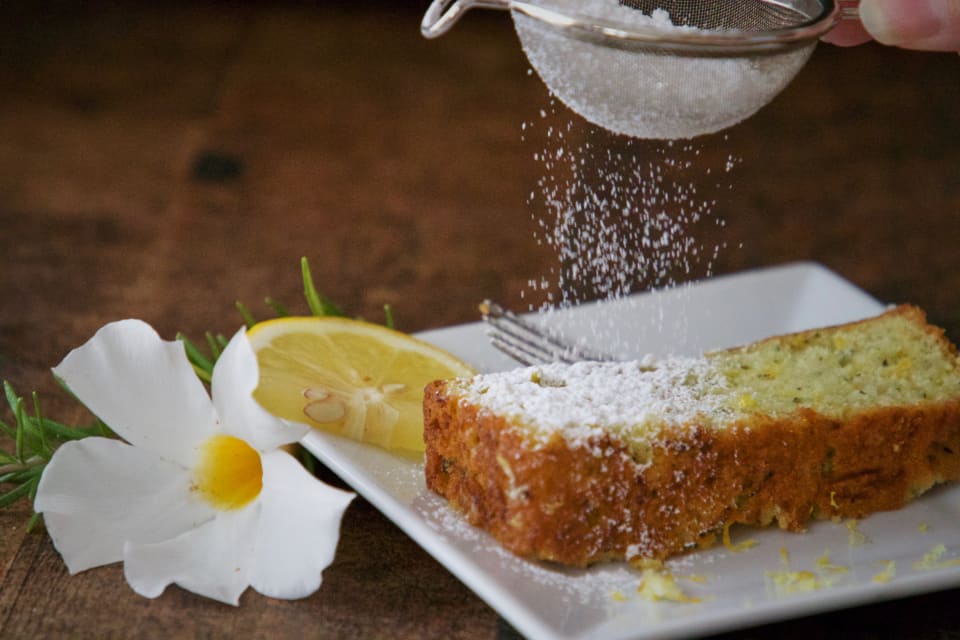 Fatia de bolo de limão recebendo açúcar de confeiteiro, com meia rodela de limão ao lado