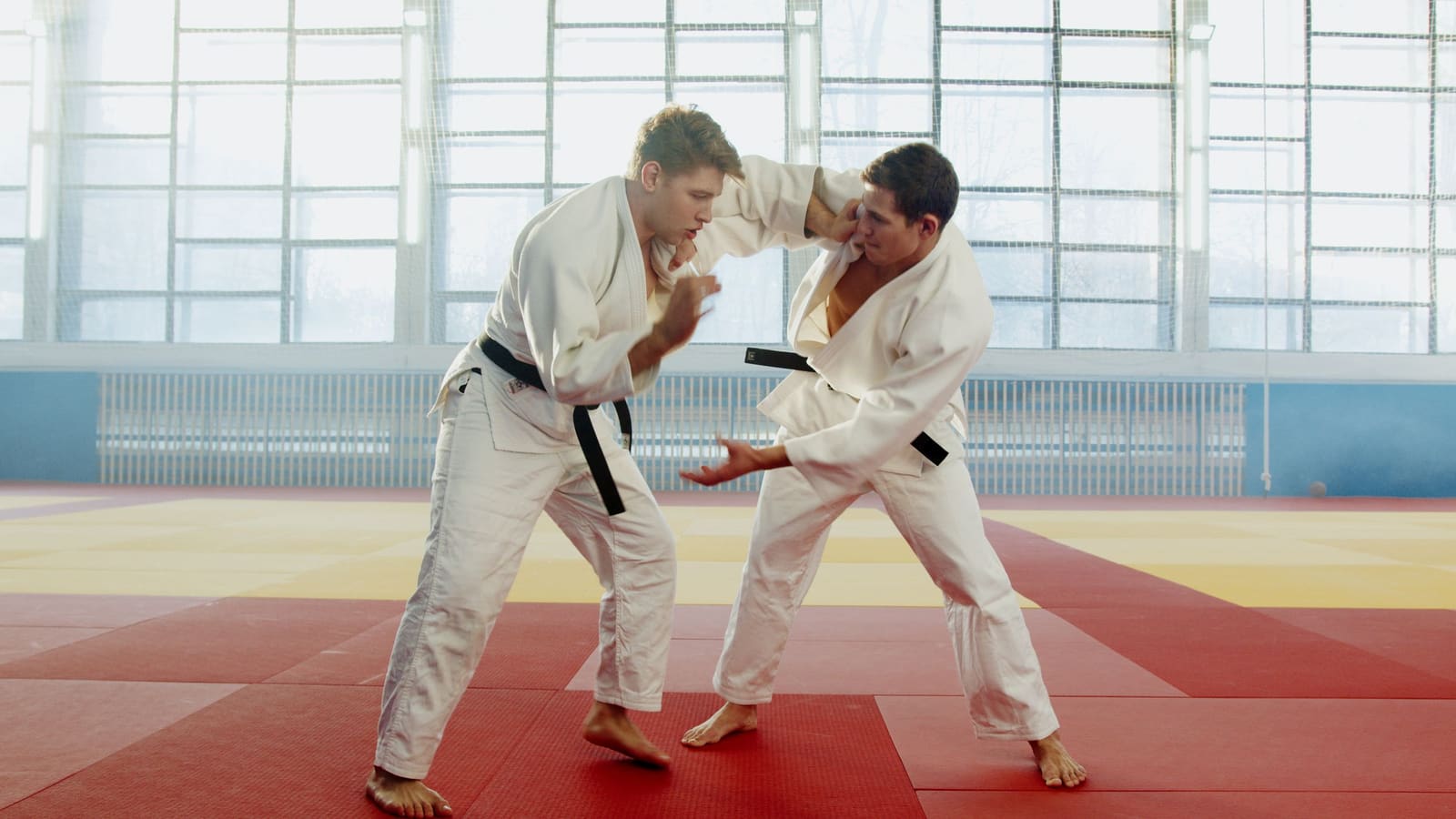 Dois judocas de kimono branco e faixa preta treinando uma pegada no tatame vermelho e amarelo de um dojo com janelas grandes