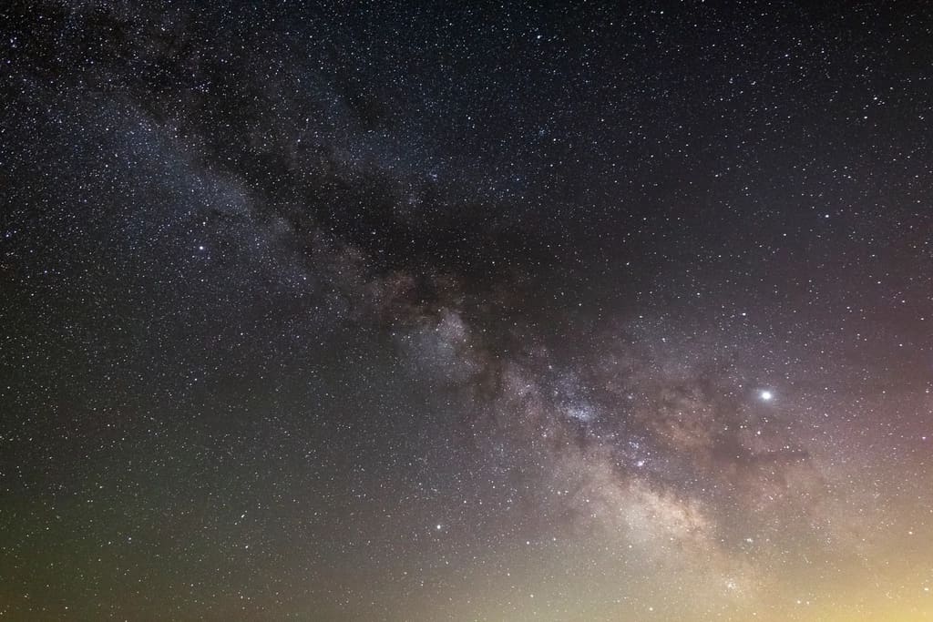 Céu noturno com a Via Láctea, cheio de estrelas, sobre uma paisagem escura
