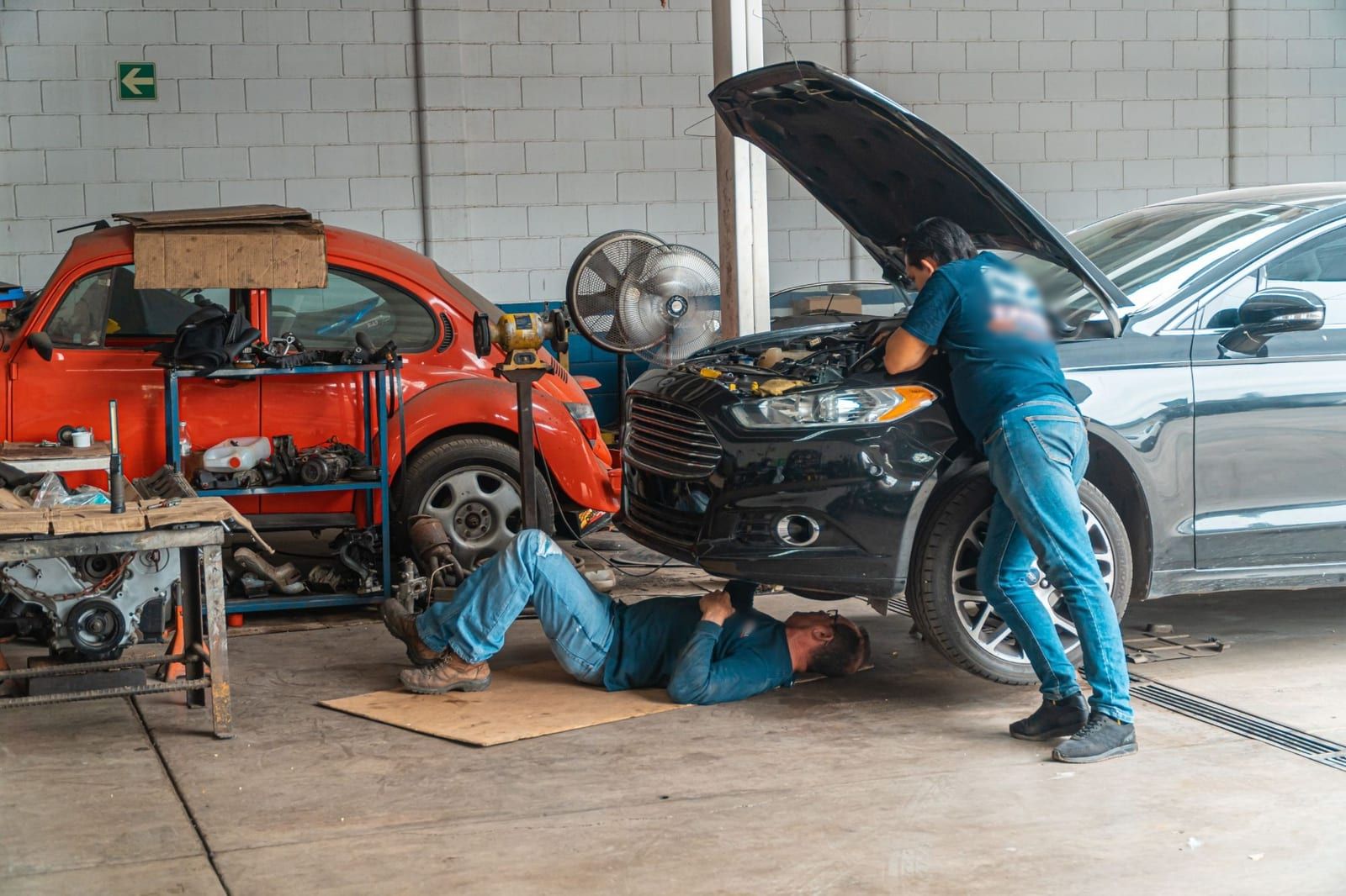 Dois mecânicos trabalhando numa oficina: um se debruça sobre o motor de um carro preto com o capô aberto e o outro está deitado no chão, embaixo do carro; ao lado, um carro laranja e uma estante com peças