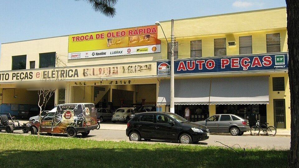 Fachada amarela de uma loja de auto peças e elétrica com uma placa azul AUTO PEÇAS e um painel de troca de óleo rápida; na rua em frente, um carro preto, uma van grafitada, um Fusca e outros carros estacionados
