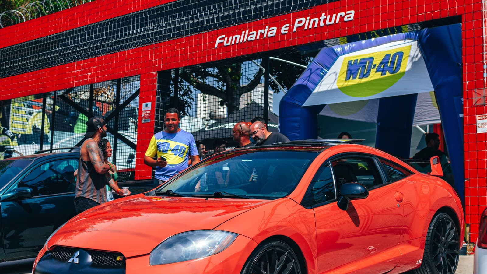 Fachada de uma funilaria revestida de azulejos vermelhos com a placa "Funilaria e Pintura"; na frente, um carro esportivo laranja e algumas pessoas conversando perto de um arco inflável azul